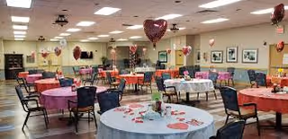 Large dining/activity room with round tables set for an event, chairs around them, and heart-shaped balloons and Valentine decorations.