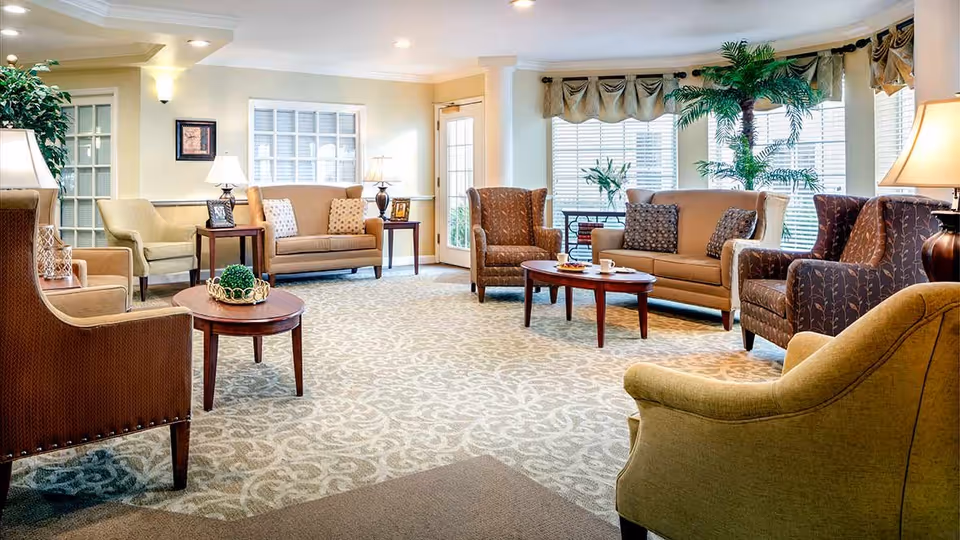 A spacious and well-lit living room area in an assisted living facility featuring multiple upholstered armchairs and sofas arranged around two wooden coffee tables. The room has large windows with blinds and valances, a patterned carpet, several table lamps, framed pictures on side tables, and a large potted plant near the windows.