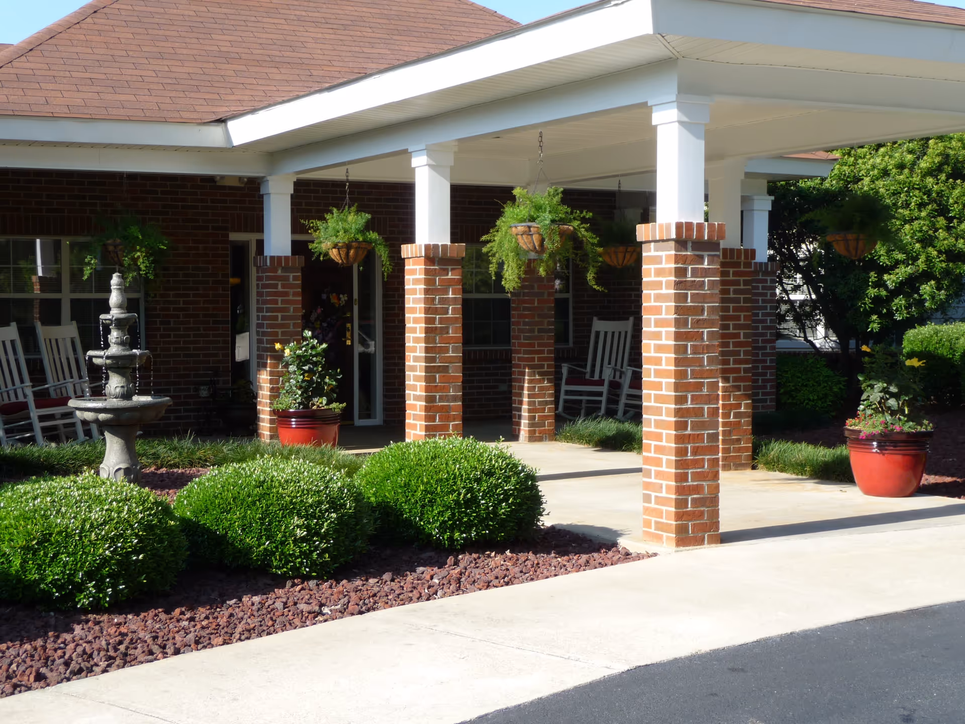 Covered entrance area of a brick building with white pillars, hanging plants, a stone water fountain, green bushes, and white rocking chairs visible inside the porch.