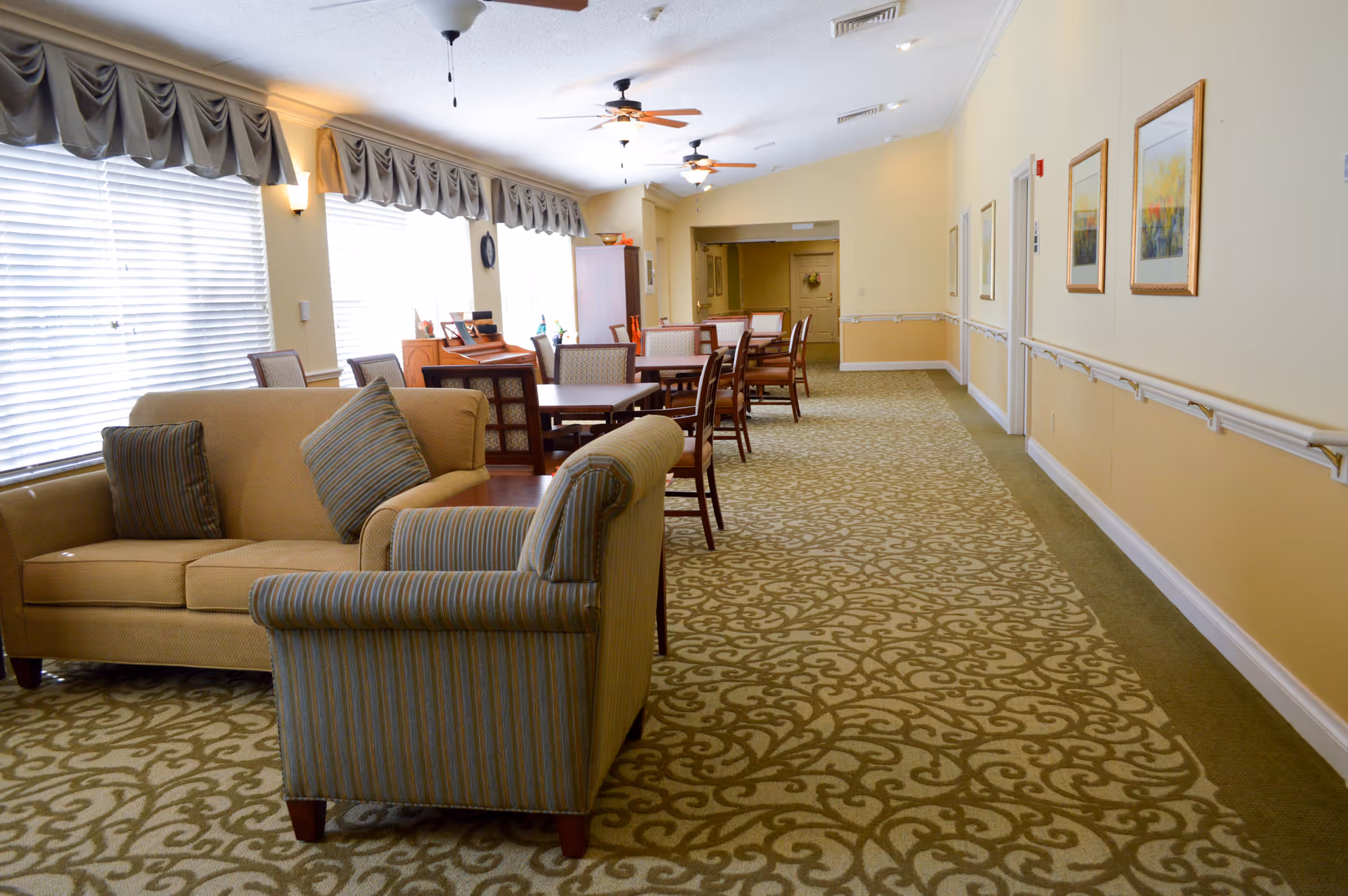 A spacious common area in a senior living facility with patterned carpet, beige walls, and multiple ceiling fans. The room features a beige sofa and a striped armchair in the foreground, several wooden tables with chairs arranged in the middle, and large windows with blinds and valances on the left side. The right wall is decorated with framed artwork and has handrails along its length.