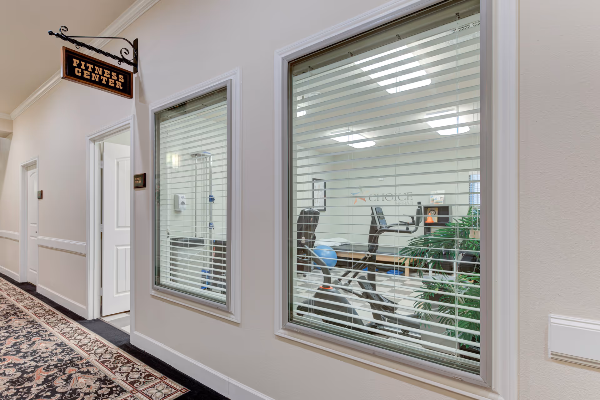 View through windows into a fitness center with exercise equipment including a stationary bike and an exercise ball, located in a hallway with a patterned carpet and a sign above the door that reads 'Fitness Center'.