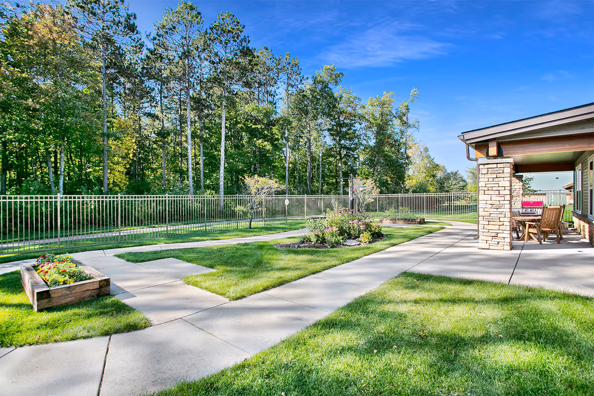 Outdoor patio and landscaped lawn with walkways, raised planters, and seating beside a building under a clear blue sky.