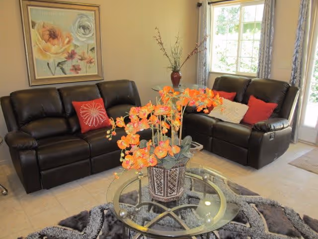 Two black leather sofas face a glass coffee table topped with an orange orchid arrangement in a bright living room with wall art and a window.