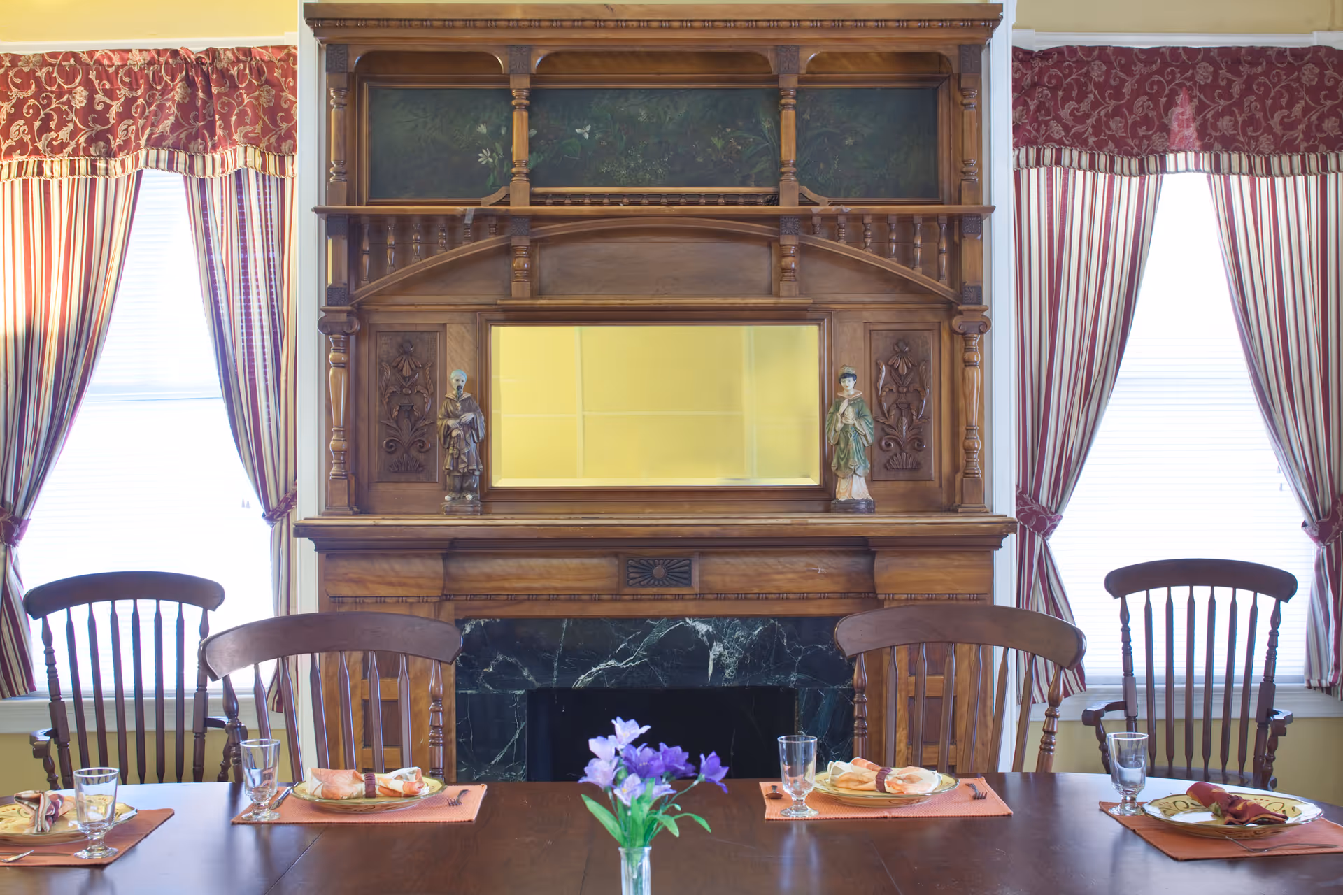 A dining room with a wooden table set for four people, each place setting having a plate with a napkin and utensils, and a glass. The table has a small vase with purple flowers in the center. Behind the table is an ornate wooden fireplace mantel with a mirror and two decorative figurines. The room has two windows with red and white striped curtains and valances.