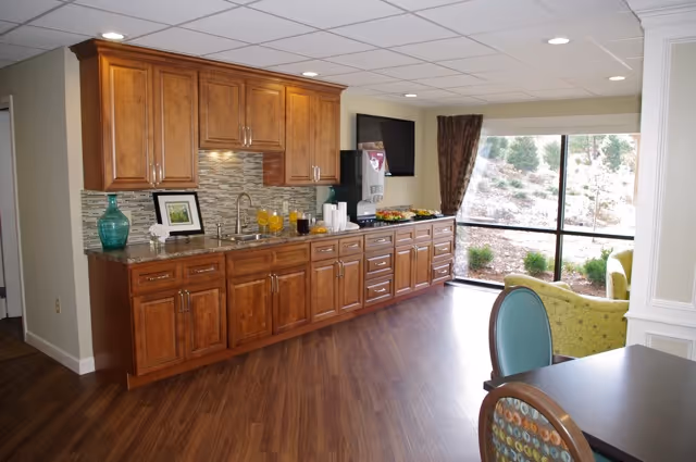 Interior view of a room with wooden cabinets and a countertop along one wall, featuring a sink, a decorative vase, framed picture, and a beverage dispenser with cups. The room has wood flooring, a large window with a view of an outdoor garden area, and some chairs and a table partially visible in the foreground.