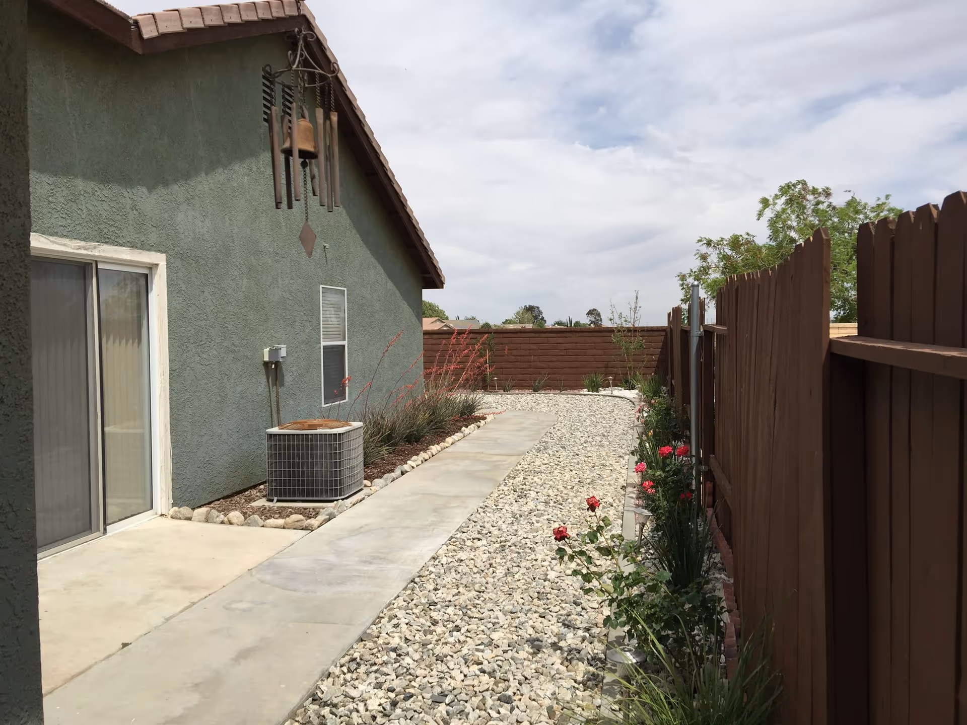 Outdoor side yard area of a building with a concrete walkway and gravel ground cover. The building has green stucco walls, a sliding glass door, a window, and an air conditioning unit. There is a brown wooden fence on the right side with some plants and flowers growing along it. The sky is partly cloudy.