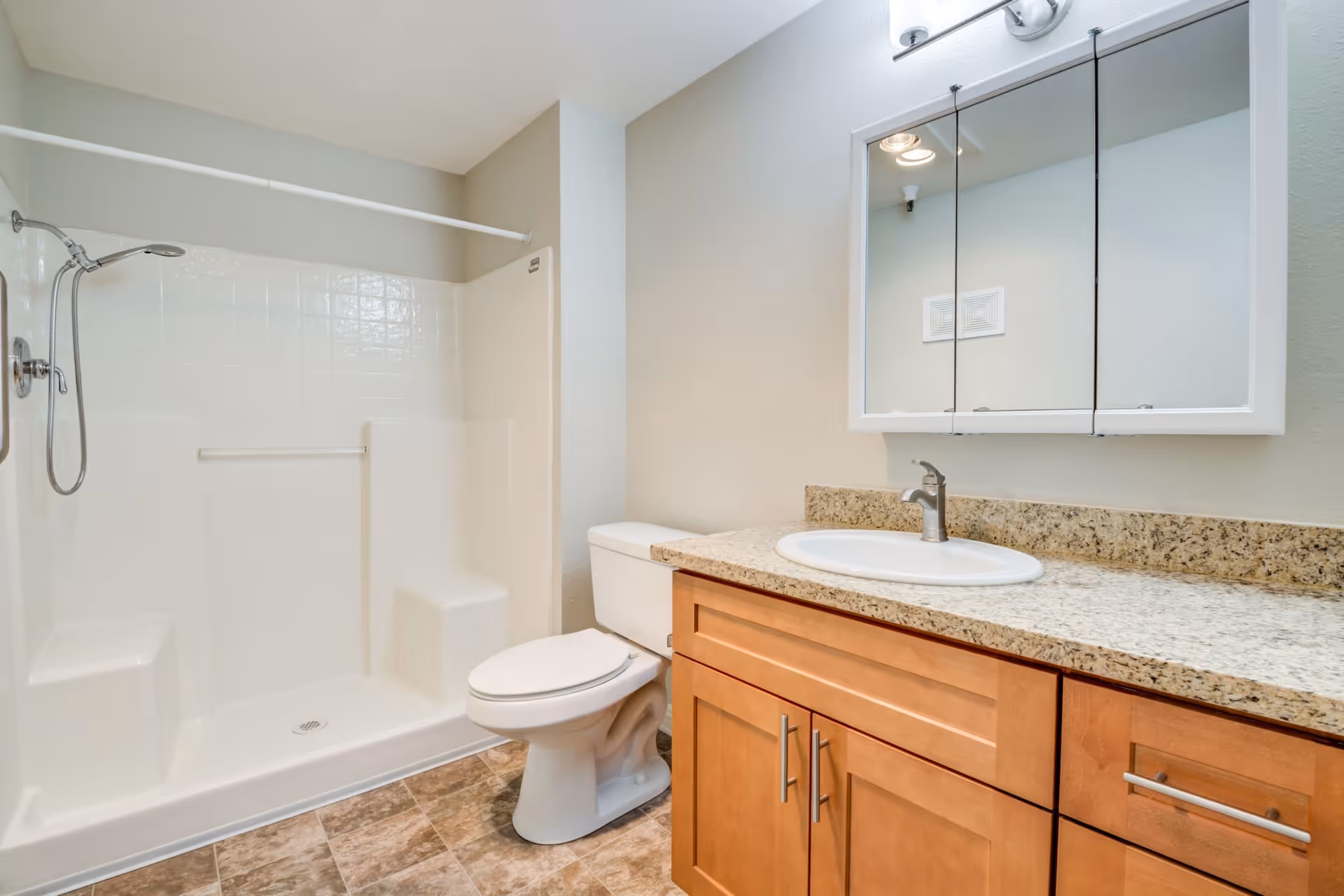 A clean bathroom featuring a walk-in shower with built-in seats and a handheld showerhead, a white toilet, and a wooden vanity with a granite countertop and an oval sink. Above the sink is a mirrored medicine cabinet with three panels.