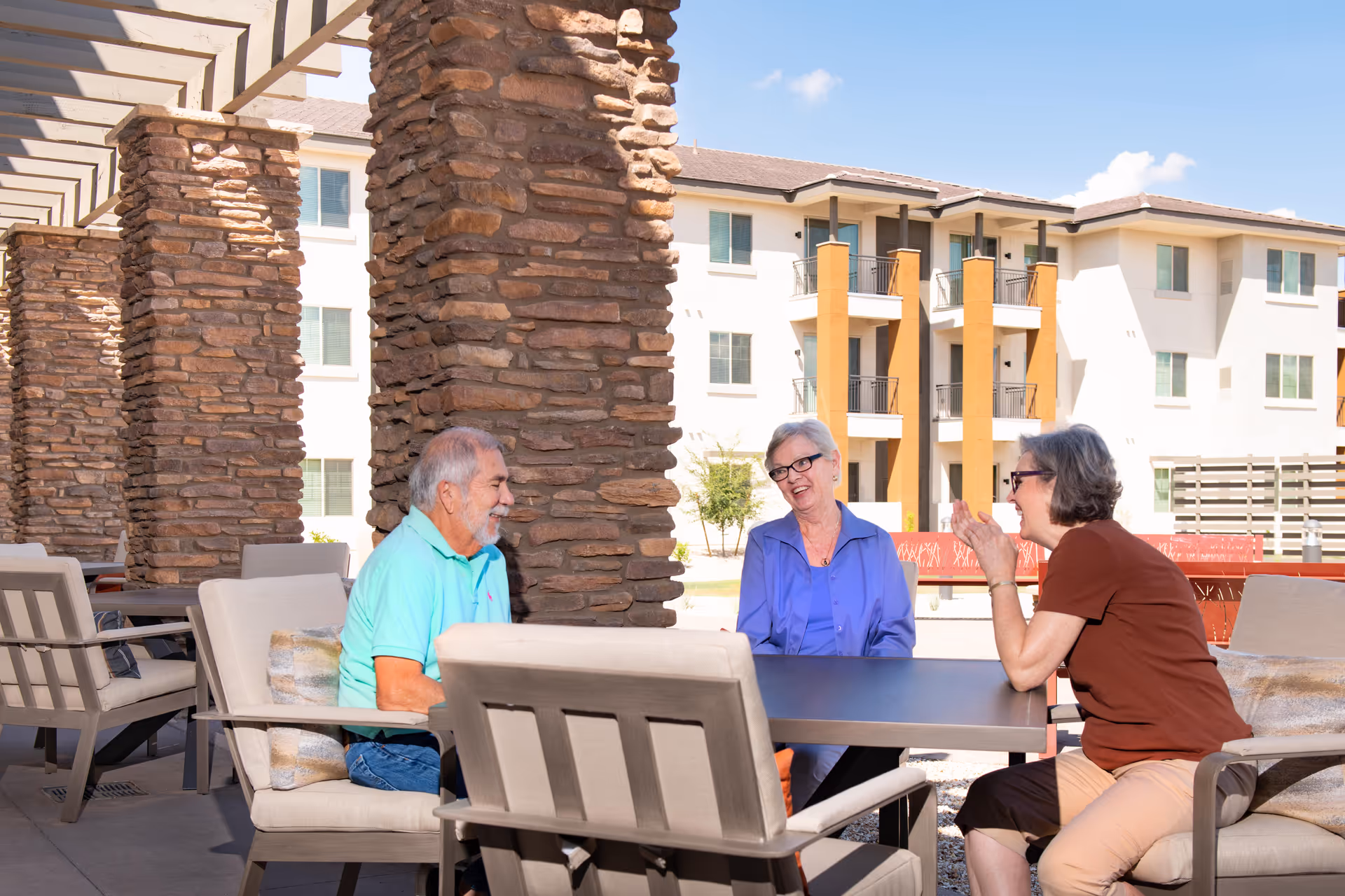 Three older adults seated and talking at a shaded outdoor patio table with a multi-story senior living building in the background.