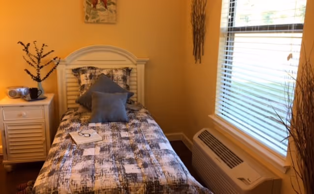 A small bedroom with a single bed covered in a patterned gray and white bedspread, two gray pillows, and a book with glasses resting on the bed. Next to the bed is a white nightstand with a small radio, a black mug, and a decorative branch arrangement. A window with white blinds is on the right wall, and an air conditioning unit is below the window. The walls are painted a warm yellow color, and there is a piece of artwork hanging above the bed.