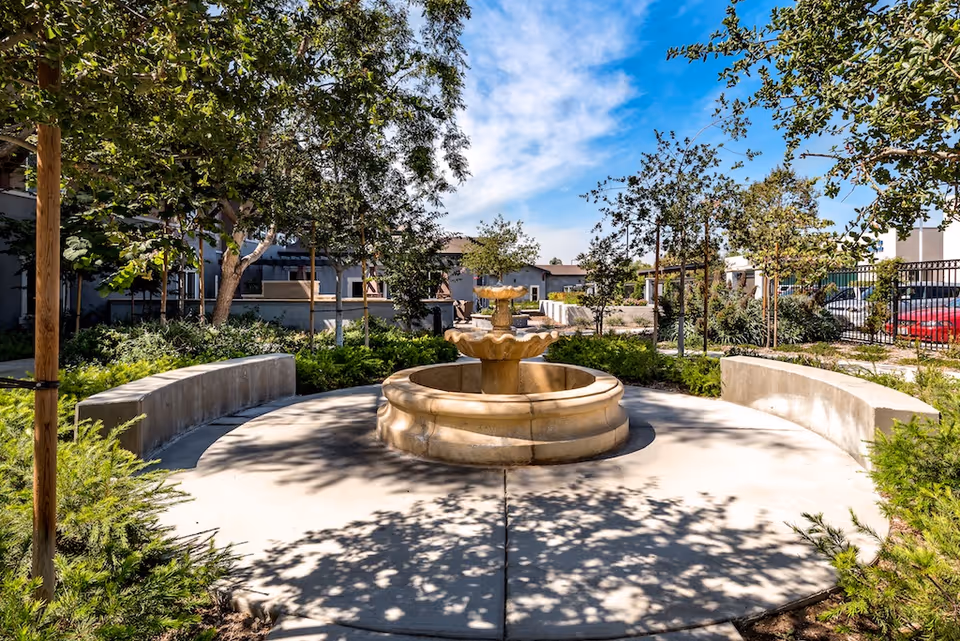 Circular stone fountain in a sunny landscaped courtyard with curved concrete benches and trees.