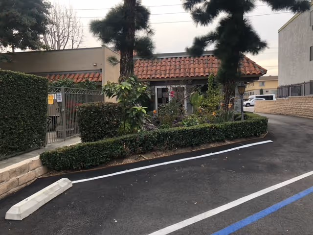 Exterior view of a building with a red tile roof and beige walls, surrounded by trees and bushes. There is a paved parking area with marked parking spaces in the foreground.