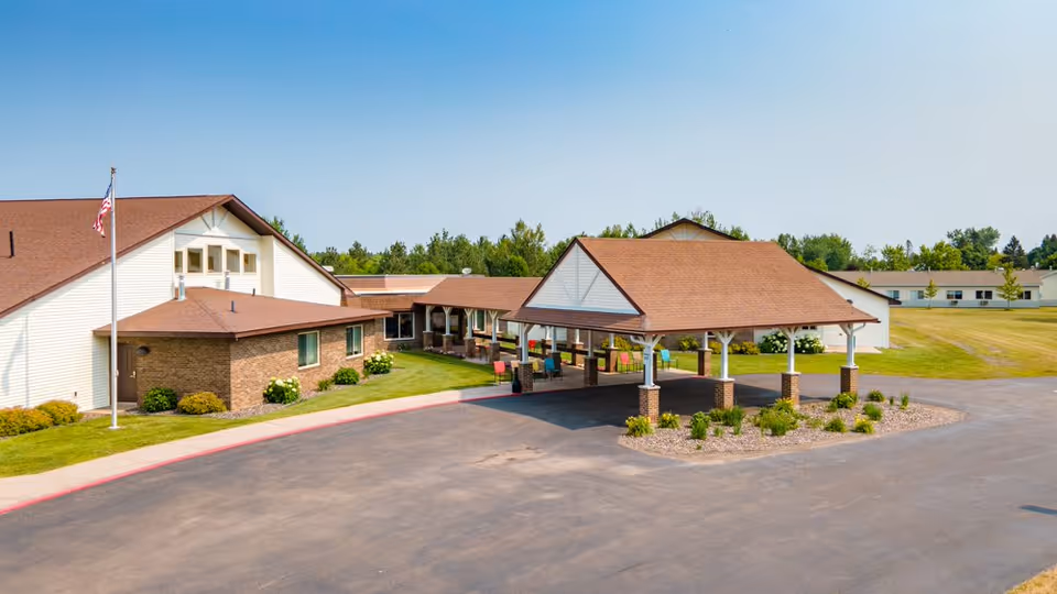 Front entrance of a single-story senior living building with a covered drop-off canopy, flagpole, and landscaped lawn.