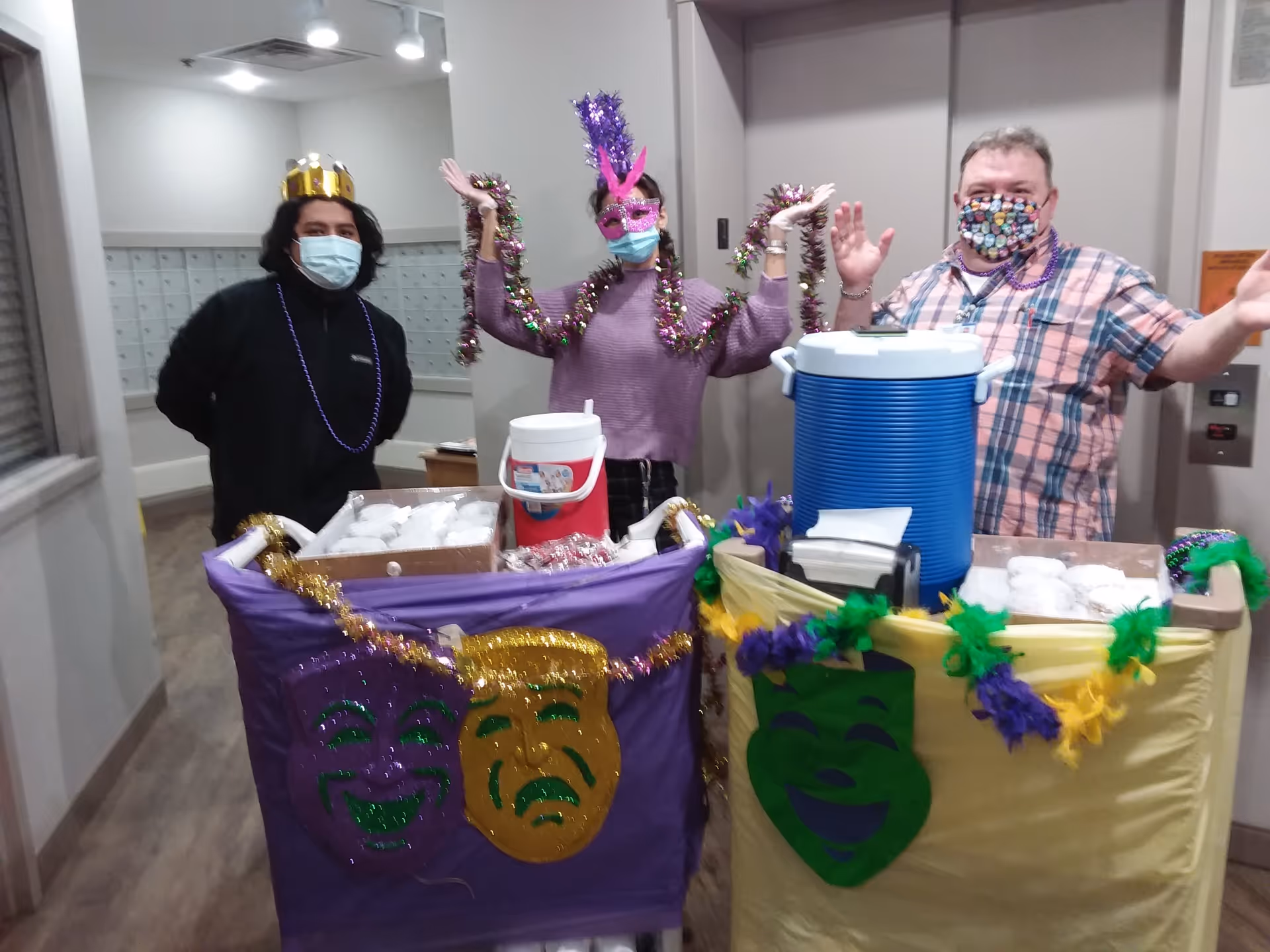 Three people wearing face masks stand behind two decorated carts filled with food and drinks. One person wears a gold crown and a black jacket, the middle person wears a purple sweater and a purple mask with feathers, and the third person wears a plaid shirt and a colorful mask. The carts are decorated with Mardi Gras-themed masks and garlands in purple, green, and gold colors. The setting appears to be an indoor hallway with mailboxes on the wall.