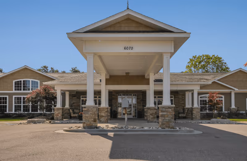Front exterior view of a single-story assisted living facility building with a covered entrance supported by four white columns with stone bases. The building has large windows, a beige and brown color scheme, and the number 6070 displayed above the entrance. There are small trees and landscaping on either side of the entrance under a clear blue sky.