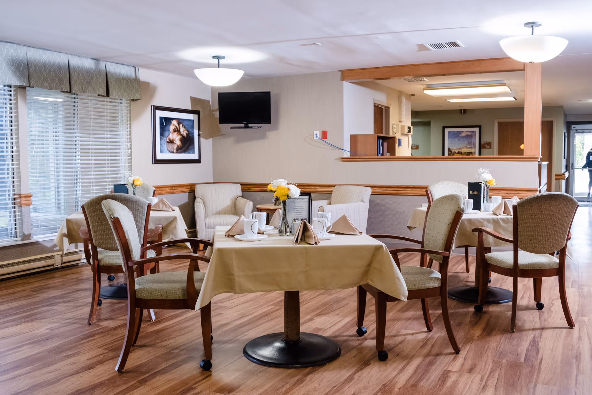 Small dining area in a senior living facility with several set tables, chairs, and floral centerpieces.
