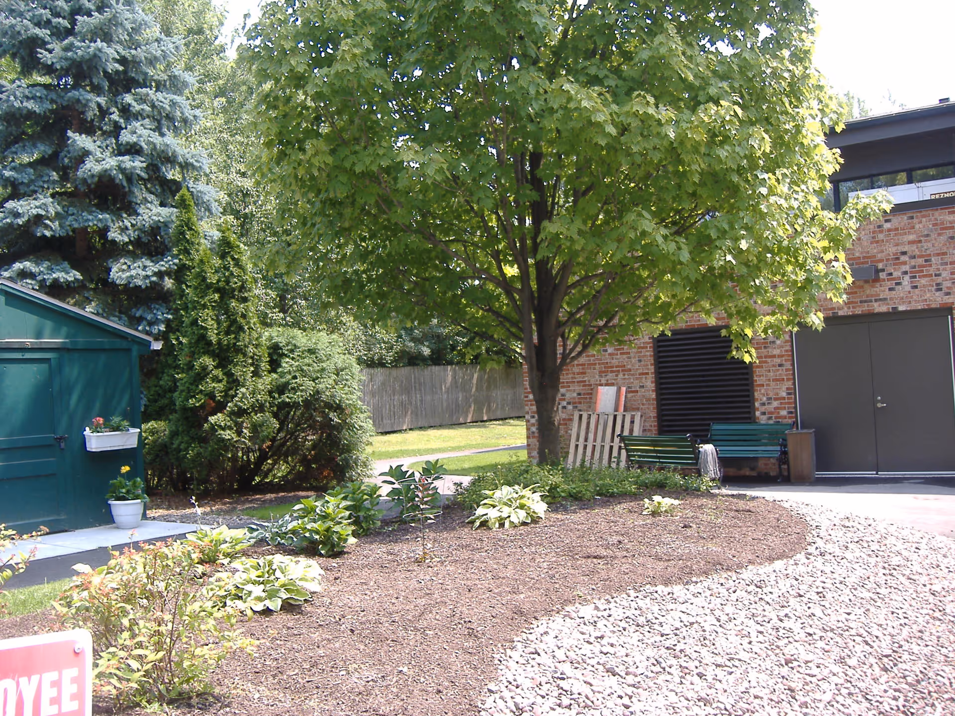 Outdoor garden area with a large leafy tree, various shrubs and plants, a green shed with flower pots, a brick building with a bench and double doors, and a gravel pathway.
