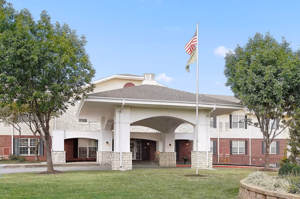 Front entrance of a two-story senior living building with a covered drive-through portico, flagpole, and trees on a lawn.