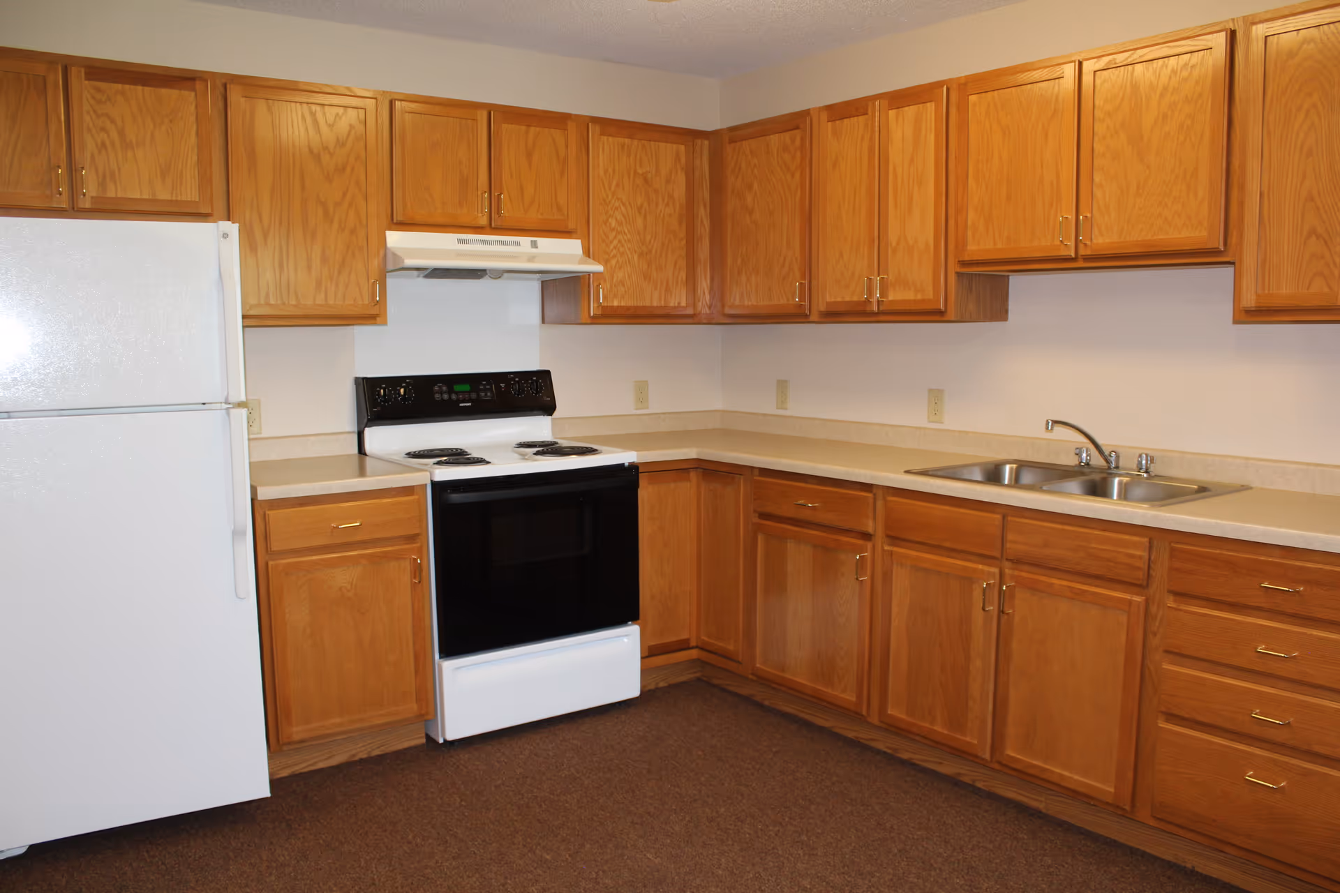 Interior view of a kitchen with wooden cabinets, a white refrigerator, a white electric stove with a range hood, and a double sink with a faucet. The countertops are beige, and the floor is carpeted in brown.