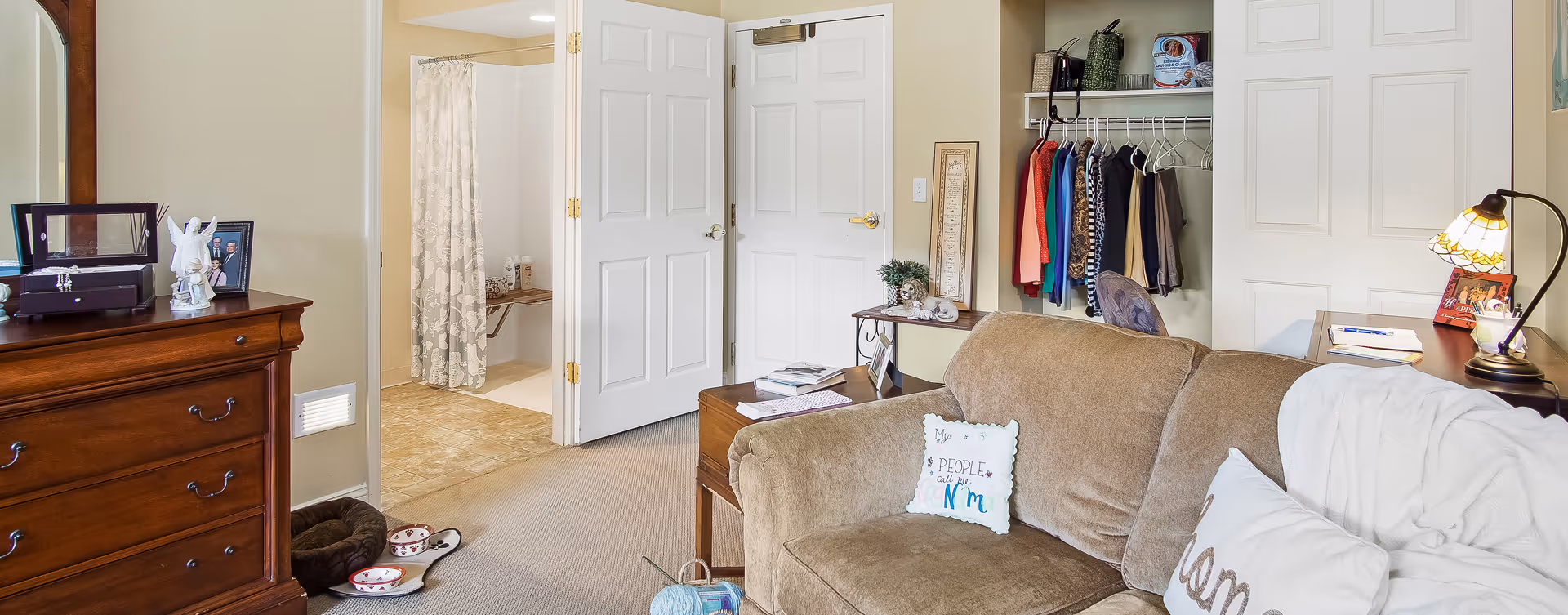 A cozy living space featuring a brown sofa with decorative pillows and a white throw blanket. To the right, there is a wooden desk with a lamp, framed photo, and notebooks. Behind the sofa, an open closet displays hanging clothes and shelves with various items. To the left, a wooden dresser with a mirror holds a jewelry box, angel figurine, and framed photo. In the background, an open door reveals a bathroom with a shower curtain and tiled floor. The room has beige walls and carpeted flooring.