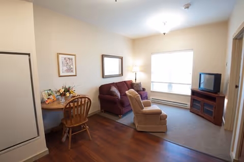 A cozy living room area with a maroon loveseat, a beige recliner chair, a wooden TV stand with an older-style television, a small round wooden table with two chairs, a lamp, and framed pictures on the wall. The room has a combination of carpet and wood flooring and a large window letting in natural light.