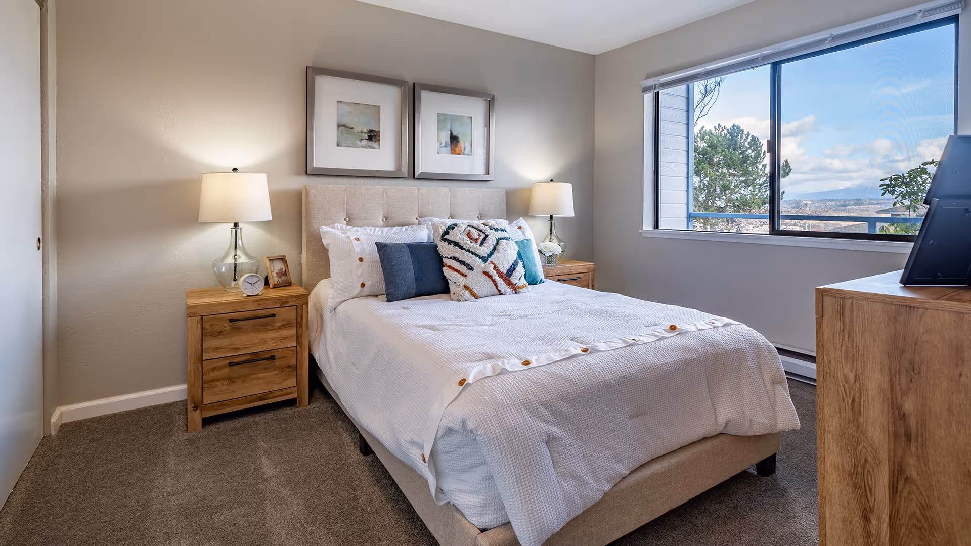 A bright and cozy bedroom with a large window showing an outdoor view. The room features a neatly made bed with white bedding and decorative pillows, two wooden nightstands each with a lamp, and framed artwork hanging above the bed. There is also a wooden dresser with a TV on top near the window.
