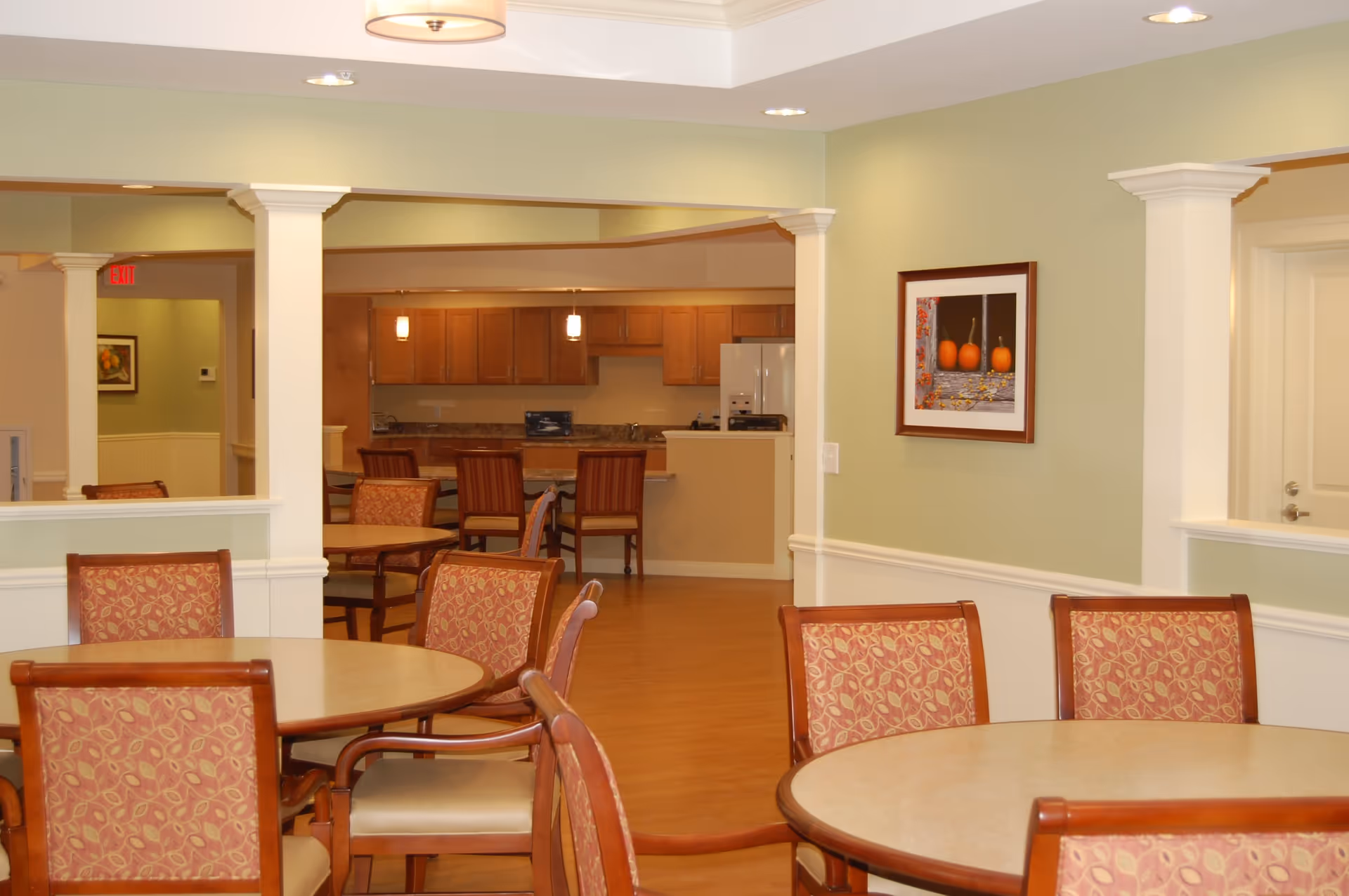 Interior view of a senior living facility dining area with round tables and chairs upholstered in a red patterned fabric. In the background, there is a kitchen area with wooden cabinets, a refrigerator, and pendant lights. The walls are painted light green with white trim, and a framed picture of pumpkins hangs on the wall.