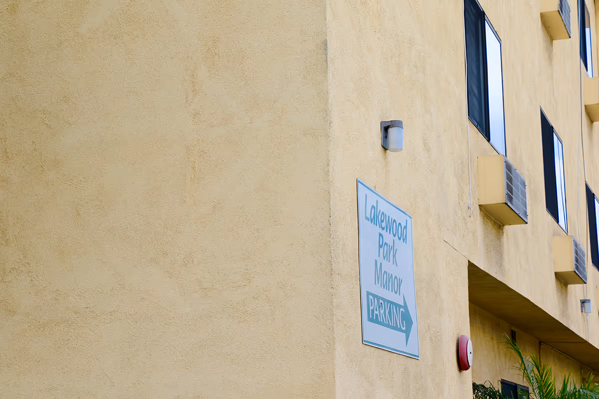 Exterior view of a beige building wall with several windows and air conditioning units. A sign on the wall reads 'Lakewood Park Manor PARKING' with an arrow pointing to the right.