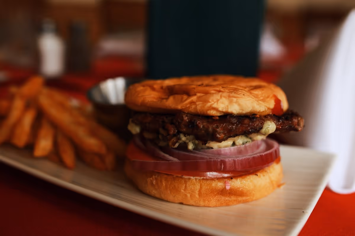Close-up of a hamburger with a beef patty, sliced red onions, and a toasted bun on a rectangular plate, served with a side of French fries and a small metal cup of dipping sauce.