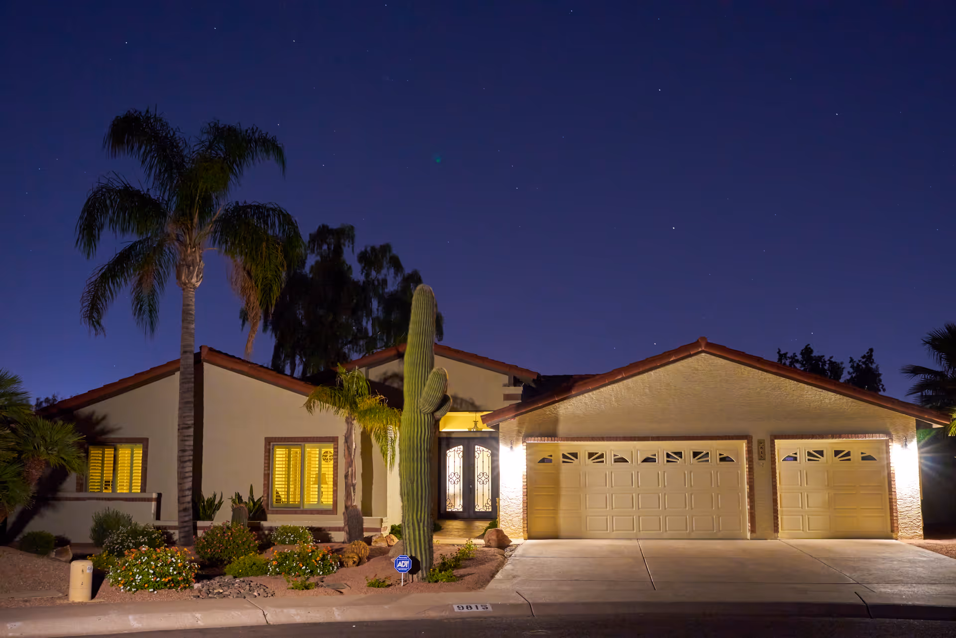 Nighttime exterior view of a single-story house with a three-car garage, illuminated by outdoor lights. The house features a tiled roof, palm trees, a large cactus, and desert landscaping with rocks and flowering plants.