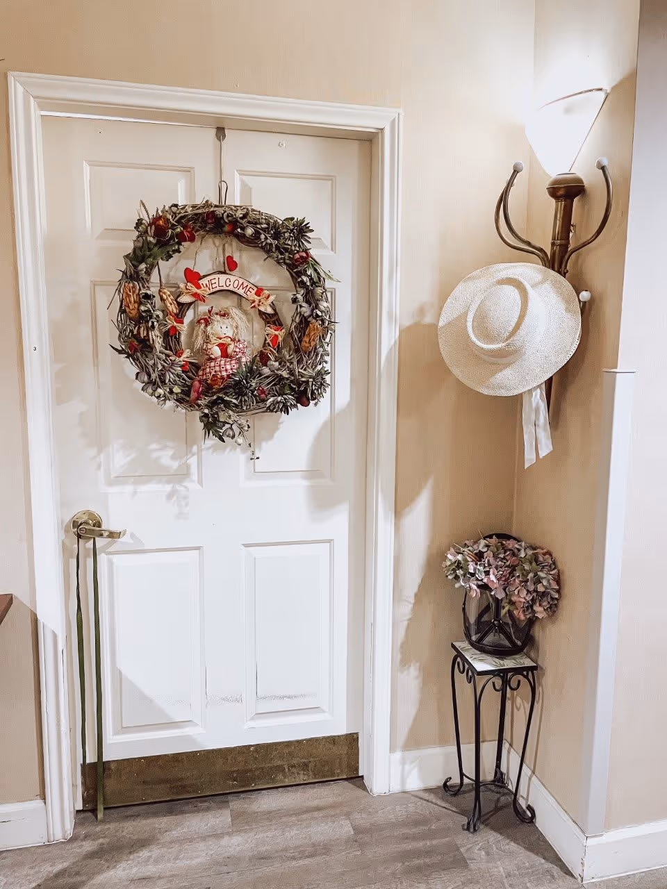 A white door decorated with a large wreath featuring a scarecrow and a 'Welcome' sign. To the right of the door, there is a wall-mounted coat rack holding a white hat, and below it is a small decorative table with a vase of flowers. The floor is wooden, and the walls are light-colored.