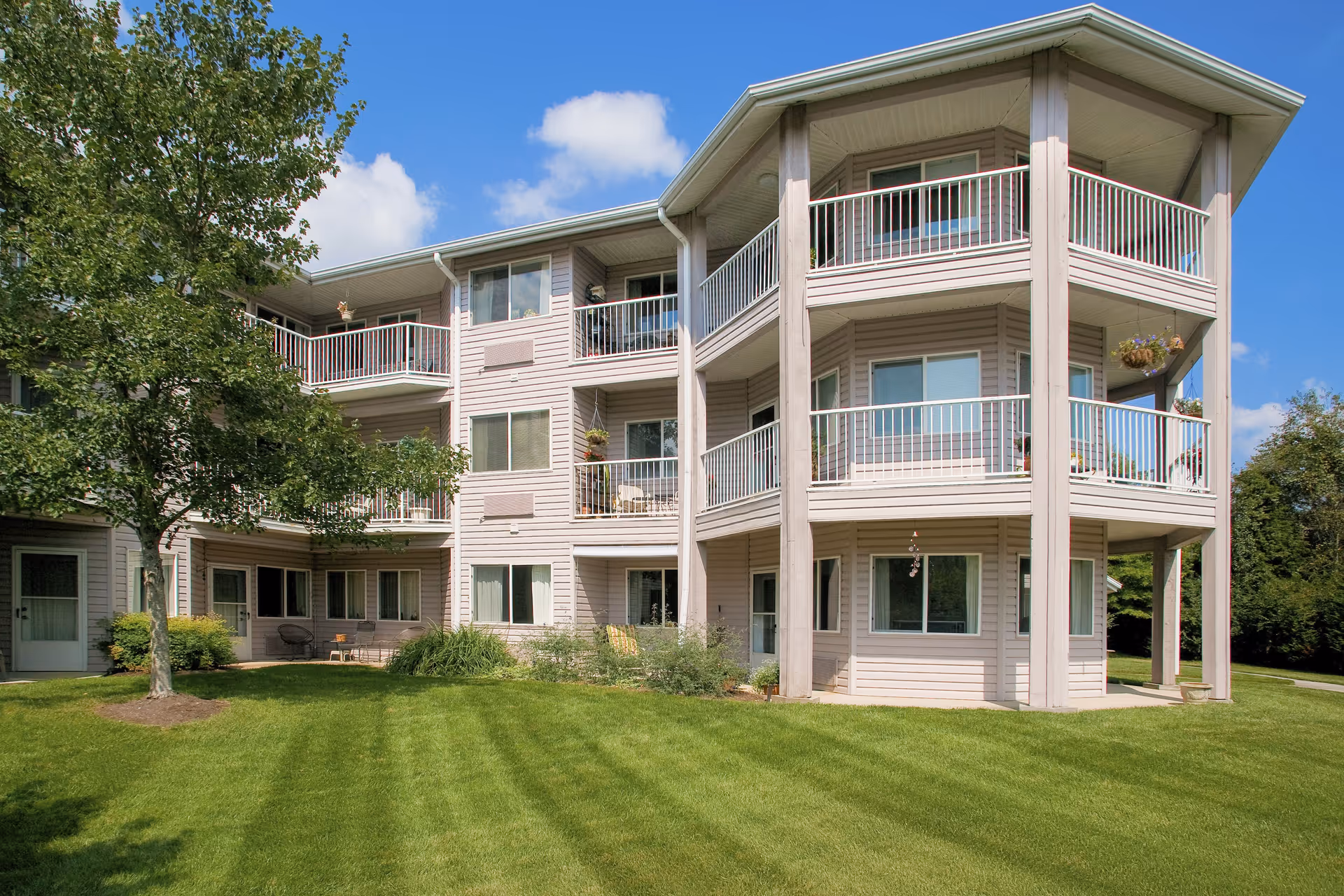 Exterior view of a three-story residential building with balconies on each floor, surrounded by a well-maintained green lawn and a tree on the left side under a blue sky with some clouds.