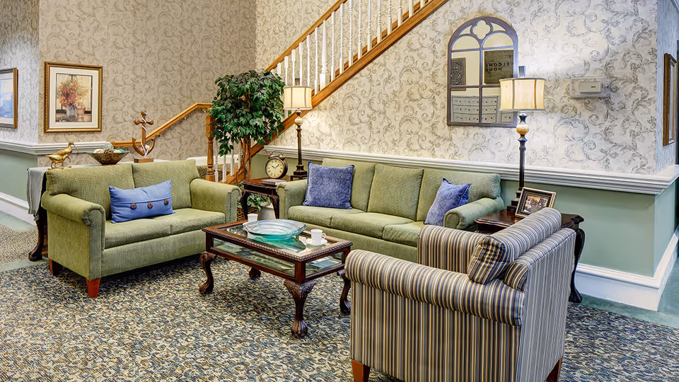 A cozy living room area with a green loveseat, a green sofa with blue cushions, and a striped armchair arranged around a wooden coffee table. The room features patterned wallpaper, a staircase with wooden railing, two table lamps, framed artwork, a decorative plant, and a wall mirror.