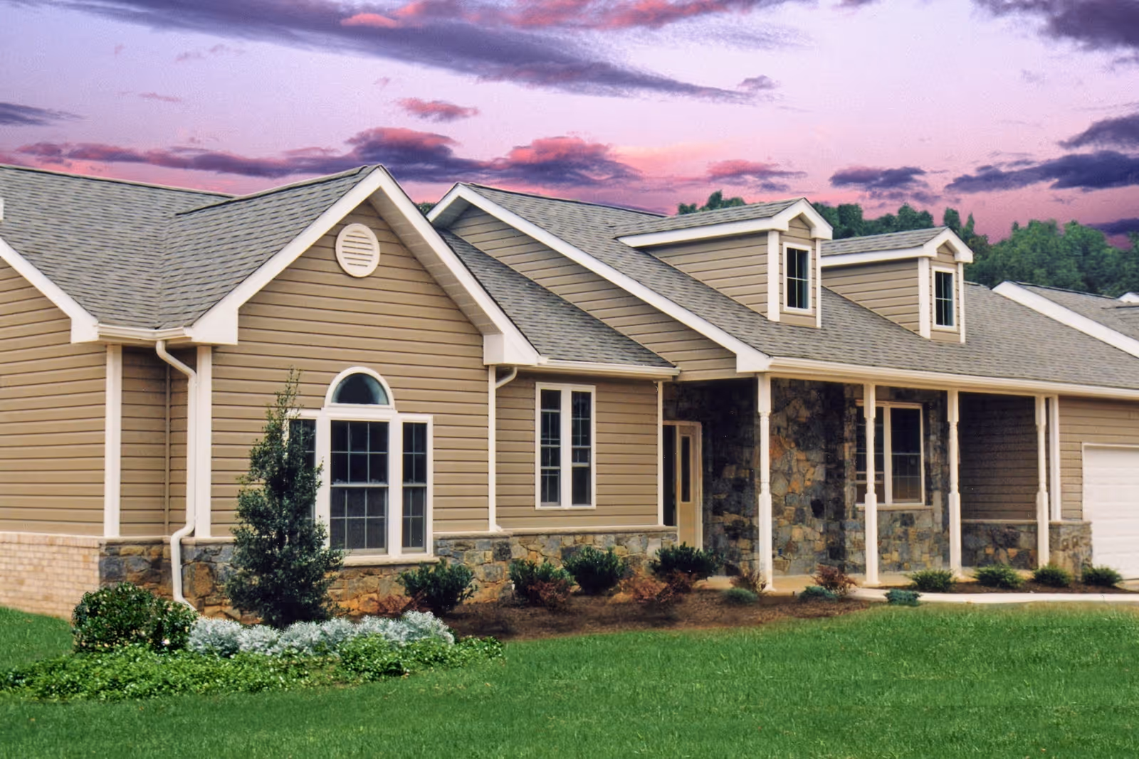 Exterior view of a single-story residential building with beige siding, stone accents, multiple windows, and a covered porch. The building is surrounded by green grass and small shrubs, with a colorful sky at sunset in the background.