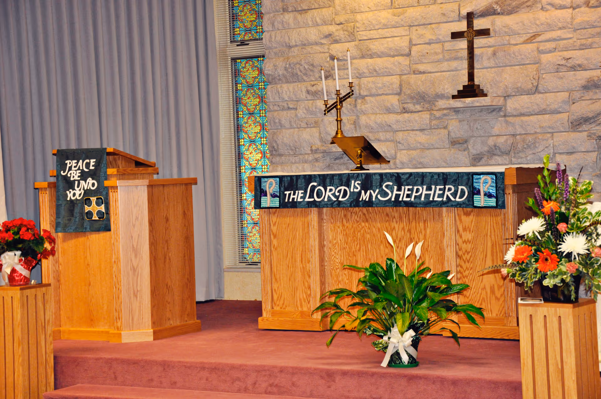 Interior view of a chapel or worship area with wooden podiums draped with cloth banners. One banner reads 'PEACE BE UNTO YOU' and another reads 'THE LORD IS MY SHEPHERD'. There are floral arrangements on either side and a stone wall with a cross mounted above the altar area. Stained glass windows are partially visible behind a curtain.