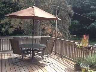 An outdoor wooden deck with a round table, chairs, and umbrella overlooking greenery.