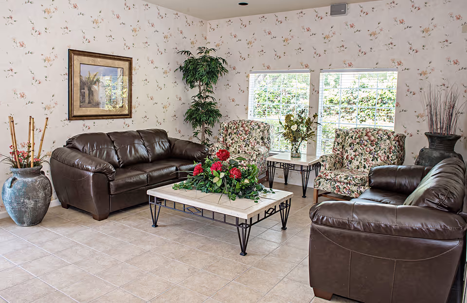 A cozy living room with two brown leather sofas and two floral-patterned armchairs arranged around a coffee table with a floral centerpiece. The room has tiled flooring, floral wallpaper, two windows with white blinds, and decorative plants in large vases.