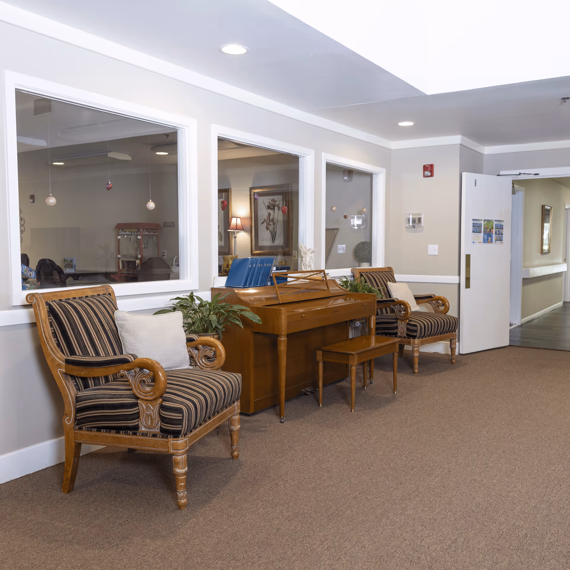 A hallway area in a senior living facility with two wooden armchairs featuring striped cushions and a wooden piano with a matching bench between them. There are plants on either side of the piano, and three large windows on the wall behind the furniture show a room with a popcorn machine and framed artwork. The hallway has beige carpet and light-colored walls with recessed ceiling lights. An open door leads to another corridor.