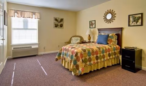 A bedroom with a single bed covered in a colorful quilt with circular patterns. The bed has a wooden headboard and is accompanied by a nightstand with a lamp and a small decorative item. There is a cushioned chair with a floral pattern next to the bed. The room has beige walls with framed butterfly artwork and a decorative round mirror above the bed. A window with blinds and a valance lets in natural light, and the floor is carpeted in a muted purple color.