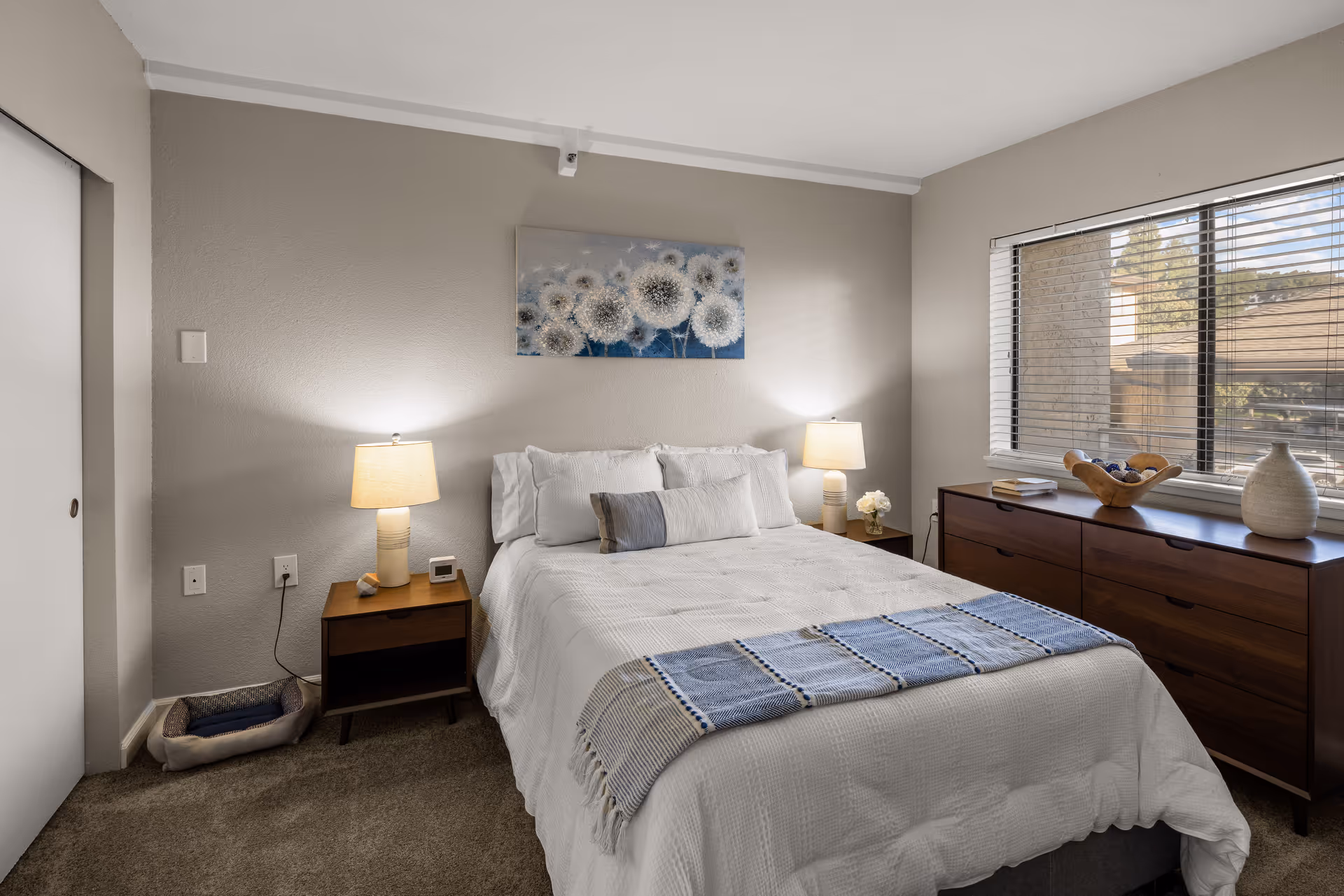 A cozy bedroom with a neatly made bed featuring white bedding and a blue throw blanket. There are two wooden nightstands on either side of the bed, each with a lamp. Above the bed hangs a painting of dandelions. A wooden dresser is positioned under a window with blinds, and decorative items are placed on top. The room has beige walls and carpeted flooring.