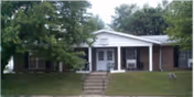 Single-story brick building with a white front porch and steps leading up to the entrance, surrounded by green grass and trees.