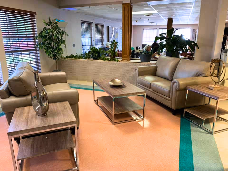 Lounge area with two leather sofas, wood-and-metal coffee and side tables, potted plants, and large windows.