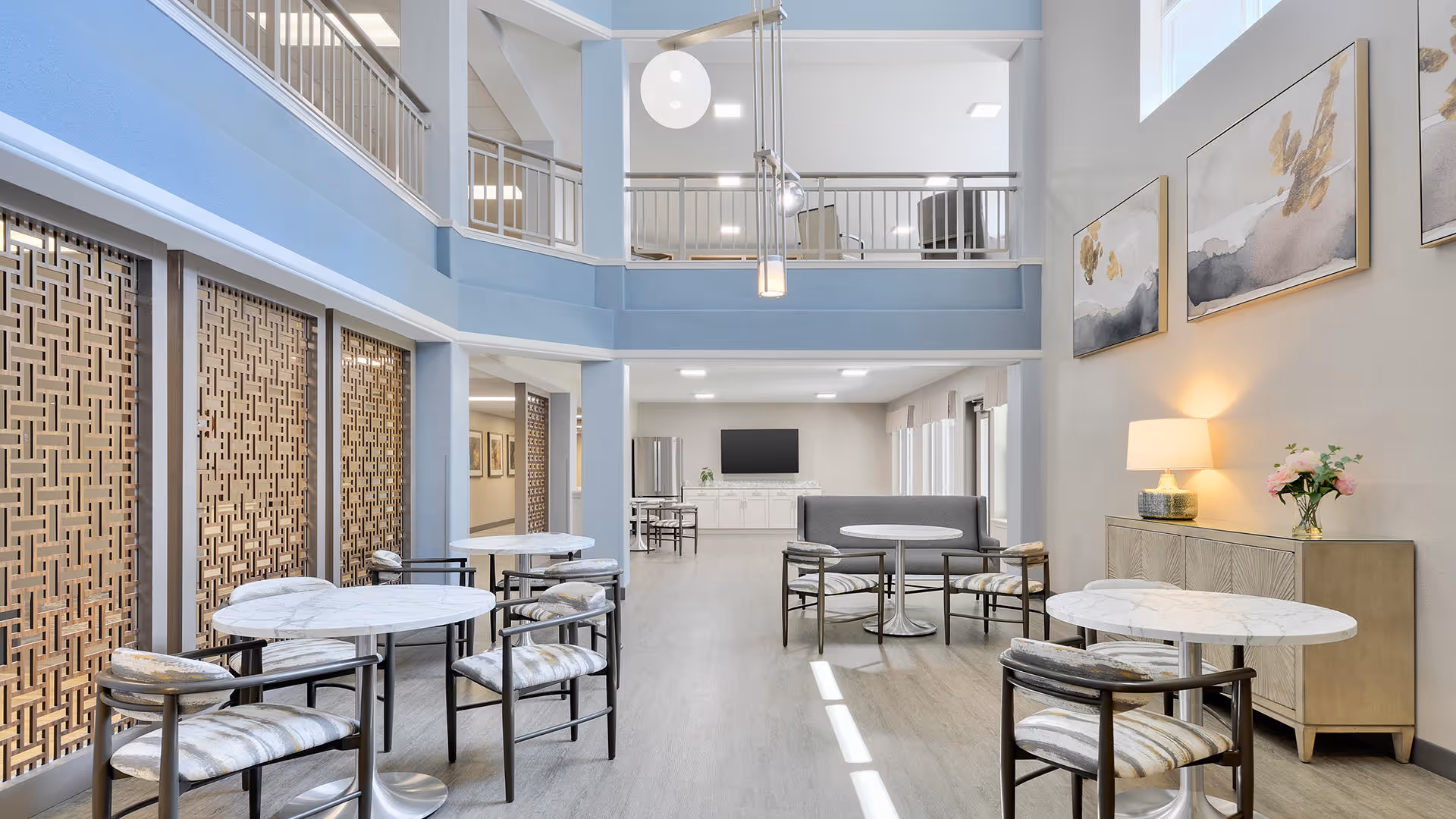 Spacious two-story communal lounge with round marble tables, upholstered chairs, a sofa, artwork, and a mezzanine balcony.