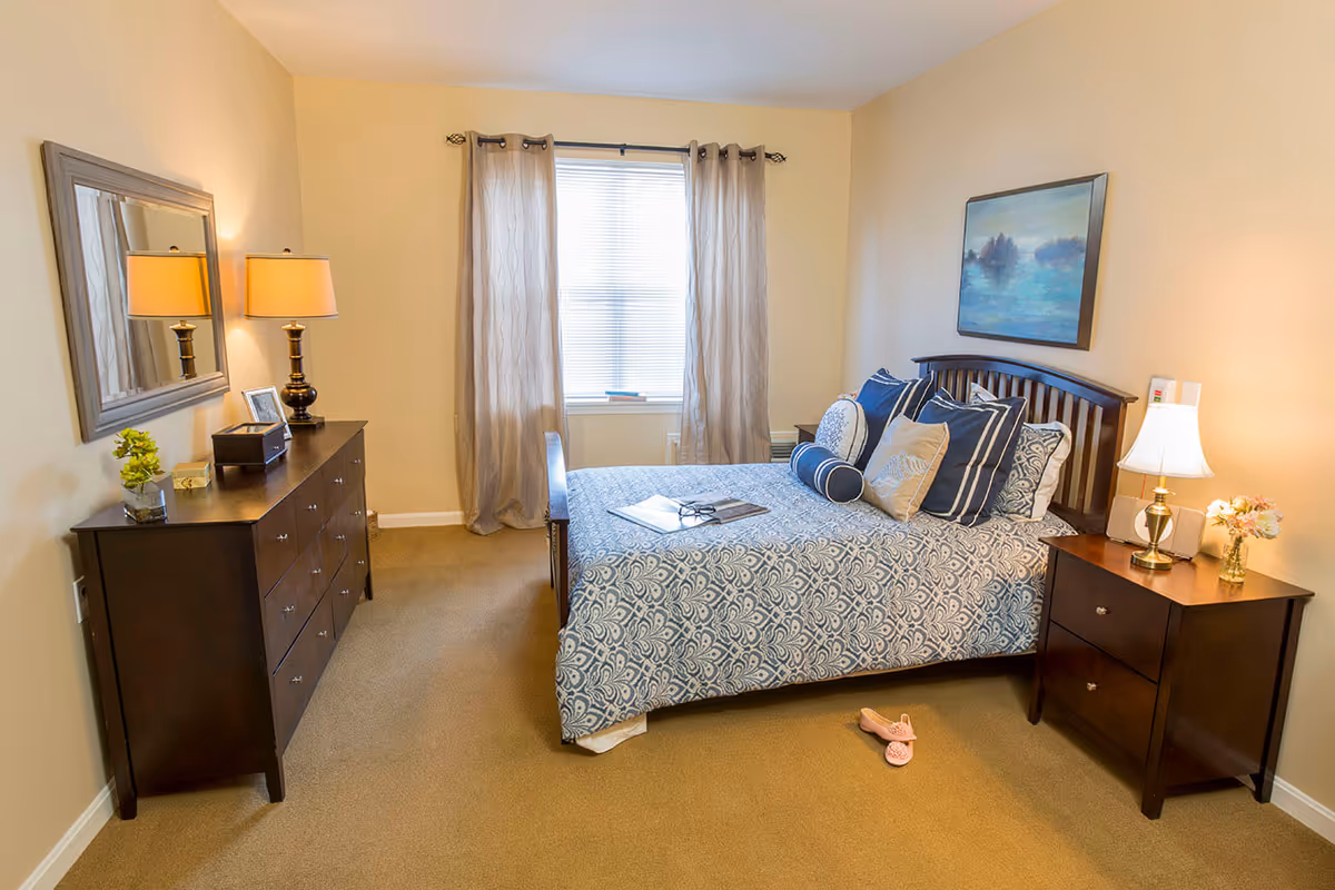 A cozy bedroom with a bed covered in a blue and white patterned bedspread and multiple pillows. There is a wooden dresser with a lamp, a mirror, and decorative items on the left side. A window with beige curtains is centered on the back wall. On the right side, there is a wooden nightstand with a lamp and a small flower vase. A pair of pink slippers is on the carpeted floor near the bed.