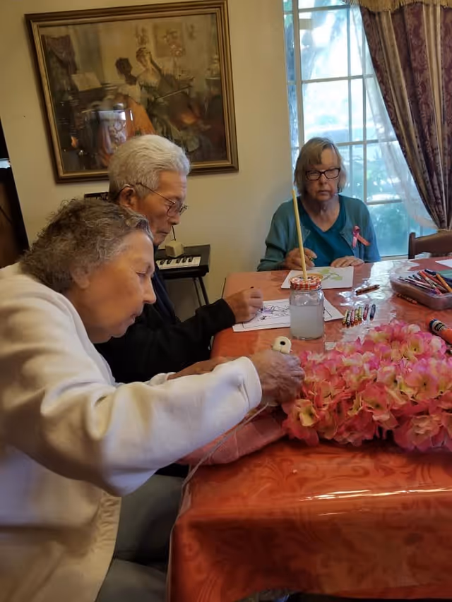 Three elderly individuals seated around a table engaged in arts and crafts activities. One person is working with pink flowers, while the other two are drawing or coloring on paper. The room has a large window with curtains and a framed painting on the wall.