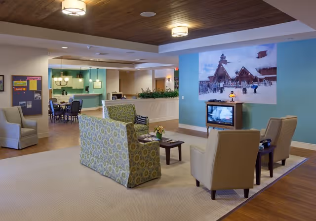 A cozy senior living facility common area with comfortable armchairs and a patterned loveseat arranged around a small coffee table. A television is placed against a blue accent wall displaying a snowy scene. In the background, there is a dining area with tables and chairs, and a kitchen with pendant lighting. The ceiling has wooden paneling and recessed lighting.