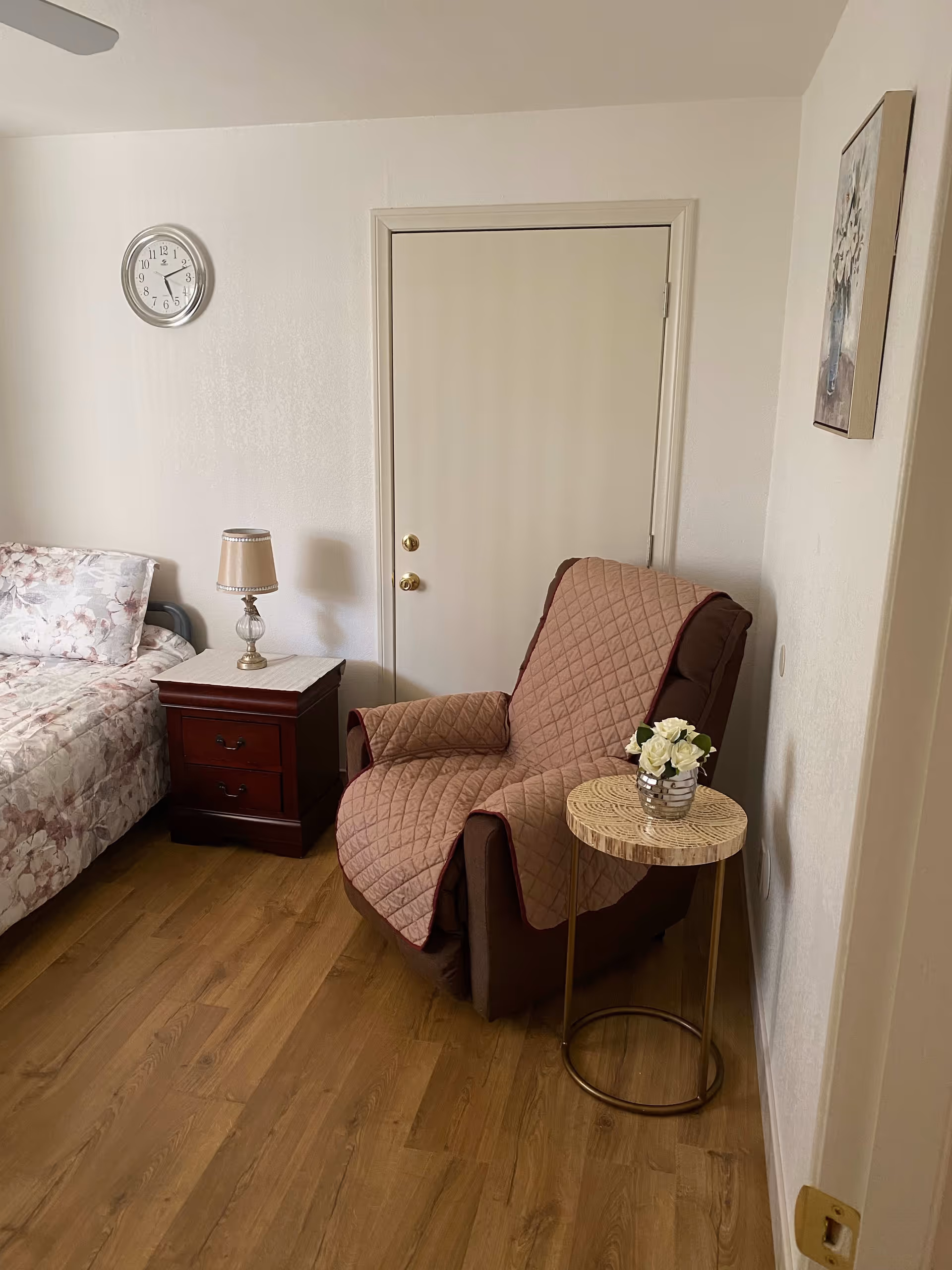 A cozy bedroom corner with a floral-patterned bed, a wooden nightstand with a lamp, a brown armchair covered with a quilted throw, and a small round side table holding a vase of white flowers. A clock is mounted on the wall above the nightstand, and a framed floral painting hangs on the adjacent wall.