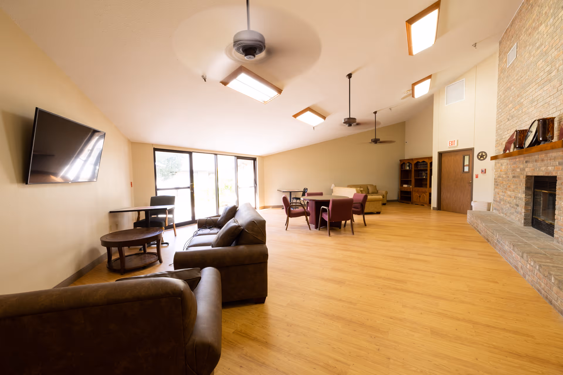 A spacious common area with wooden flooring, beige walls, and a high ceiling with ceiling fans and rectangular lights. The room features a wall-mounted flat-screen TV, several brown leather sofas, a round wooden coffee table, a few tables with chairs, a brick fireplace with decorative items on the mantel, and large windows letting in natural light.