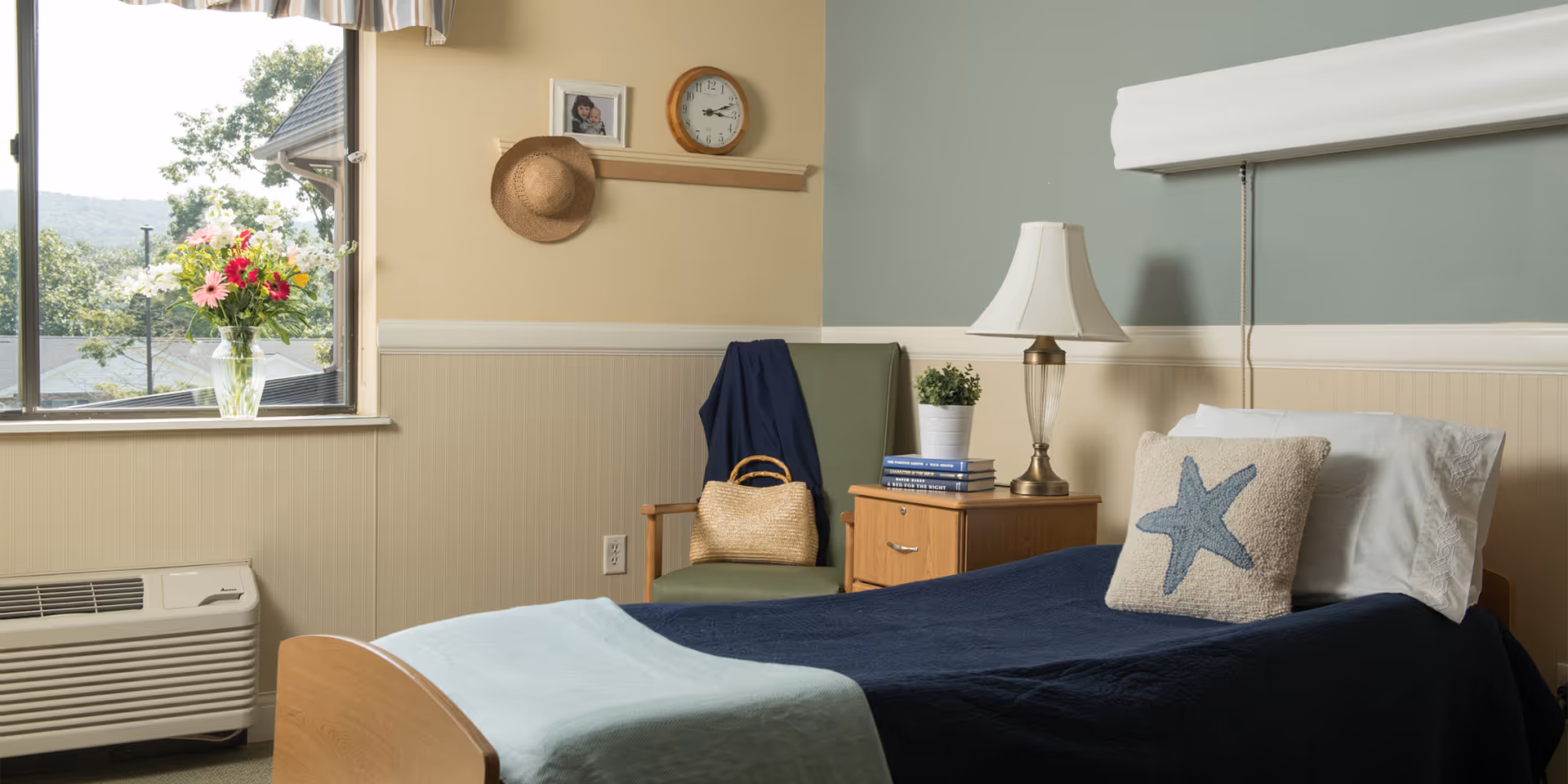 A cozy bedroom in a retirement community featuring a single bed with a navy blue blanket and a pillow with a starfish design. Next to the bed is a wooden nightstand with a lamp, a small potted plant, and a stack of books. A green chair with a woven bag and a dark blue jacket is positioned against the wall. Above the chair, a clock, a framed photo, and a hanging straw hat are displayed on a wooden rail. A window shows a view of trees and hills outside, with a vase of colorful flowers on the windowsill.