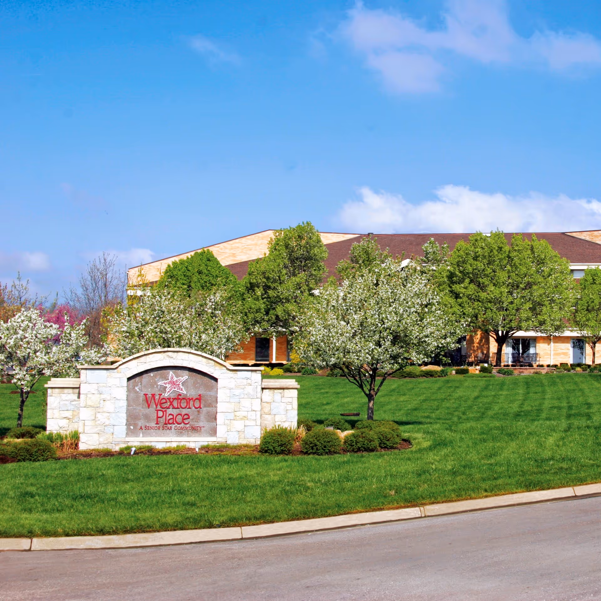 Stone sign for Wexford Place, a Senior Star community, surrounded by green grass, bushes, and blooming trees with a building and blue sky in the background.
