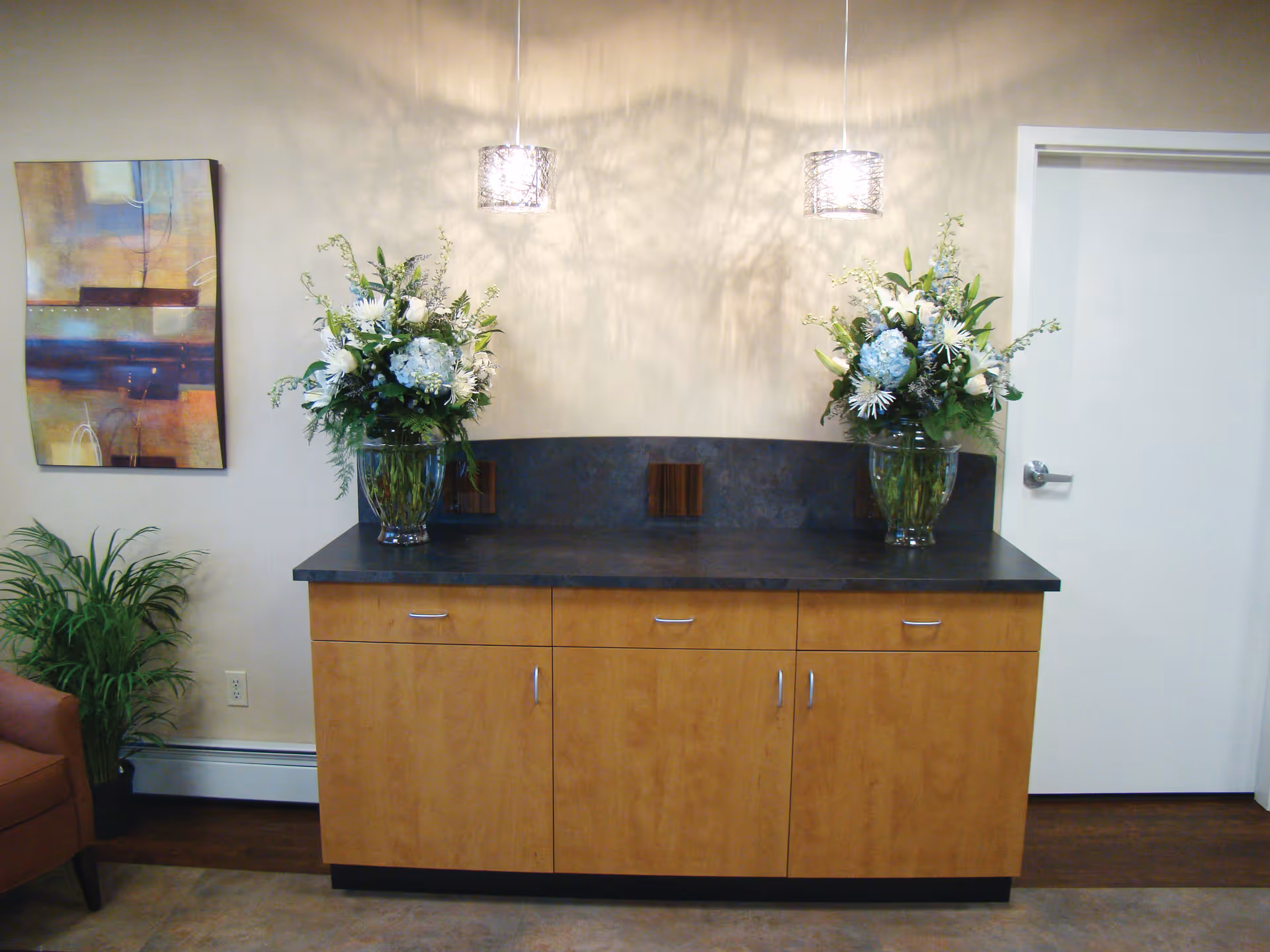 Interior view of a room with a wooden cabinet topped with a dark countertop, two large floral arrangements in glass vases, two hanging pendant lights casting shadows on the wall, a colorful abstract painting on the left wall, a green potted plant, and a closed white door on the right.